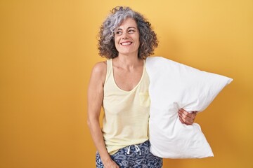 Middle age woman with grey hair wearing pijama hugging pillow smiling looking to the side and staring away thinking.