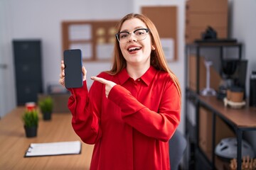 Redhead woman working at the office showing smartphone screen smiling happy pointing with hand and finger