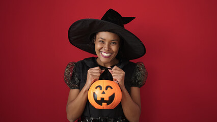 African american woman wearing witch costume holding halloween pumpkin basket over isolated red background