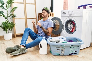 Young hispanic man putting dirty laundry into washing machine pointing to the back behind with hand and thumbs up, smiling confident