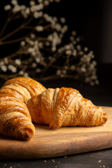 Close up of golden croissants on wooden board and dark table, with defocused white flowers and black background, vertical, with copy space