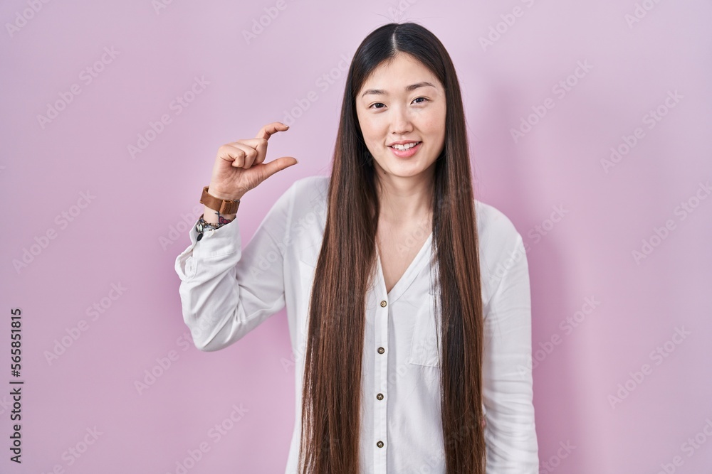 Sticker Chinese young woman standing over pink background smiling and confident gesturing with hand doing small size sign with fingers looking and the camera. measure concept.