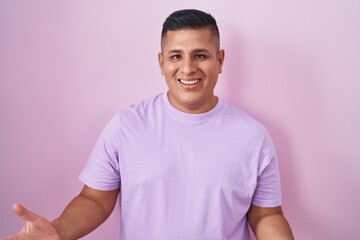Young hispanic man standing over pink background smiling cheerful with open arms as friendly welcome, positive and confident greetings