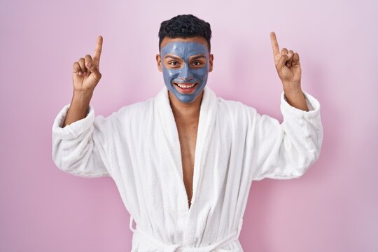 Young hispanic man wearing beauty face mask and bath robe smiling amazed and surprised and pointing up with fingers and raised arms.