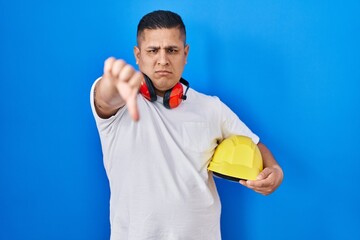 Hispanic young man holding hardhat with angry face, negative sign showing dislike with thumbs down, rejection concept