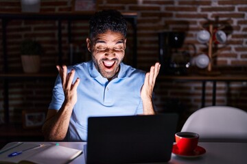 Fototapeta na wymiar Hispanic man with beard using laptop at night celebrating mad and crazy for success with arms raised and closed eyes screaming excited. winner concept