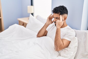 Young hispanic man suffering for headache sitting on bed at bedroom