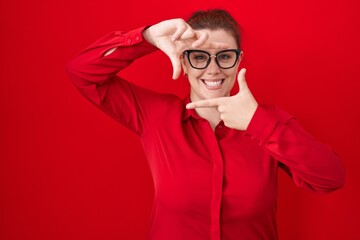 Young hispanic woman with red hair standing over red background smiling making frame with hands and fingers with happy face. creativity and photography concept.