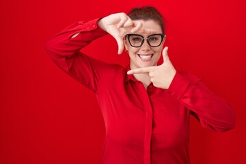 Obraz premium Young hispanic woman with red hair standing over red background smiling making frame with hands and fingers with happy face. creativity and photography concept.