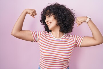 Young middle east woman standing over pink background showing arms muscles smiling proud. fitness concept.