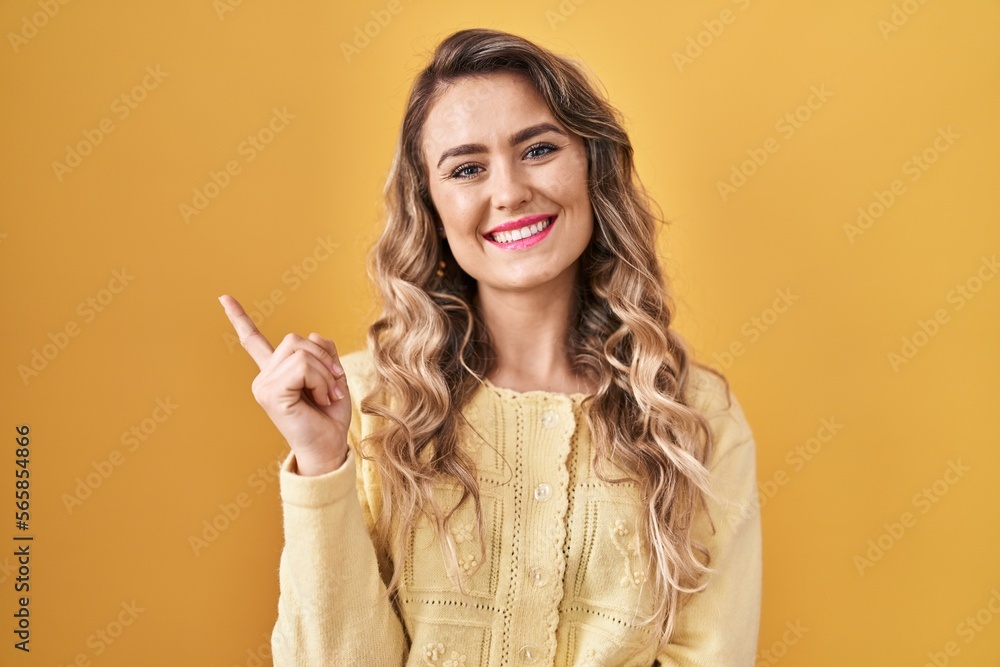 Sticker Young caucasian woman standing over yellow background with a big smile on face, pointing with hand and finger to the side looking at the camera.