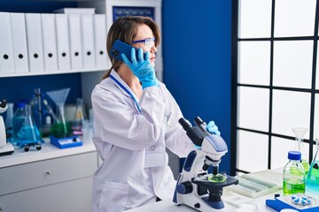 Young woman scientist using microscope talking on smartphone at laboratory