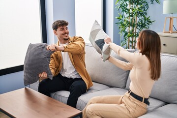 Mand and woman couple fighting with pillow sitting on sofa at home