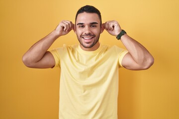 Young hispanic man standing over yellow background smiling pulling ears with fingers, funny...