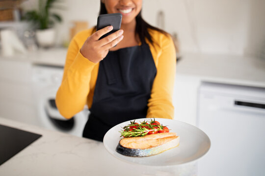 Cheerful millennial african american lady in apron taking photo of dish of fish on smartphone