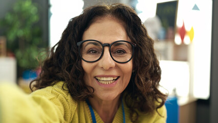 Middle age hispanic woman preschool teacher make selfie by camera sitting on chair at kindergarten
