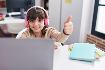 Adorable hispanic girl student using laptop and headphones doing ok gesture at classroom