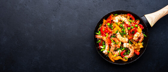 Stir fry with shrimps, red and yellow paprika, green pea, chives and sesame seeds in frying pan. Asian cuisine dish. Black stone kitchen table background, top view banner