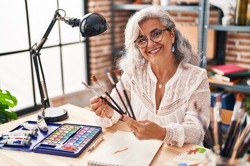 Fototapeta premium Middle age woman artist smiling confident holding paintbrushes at art studio