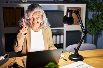 Middle age woman with grey hair working using computer laptop late at night pointing finger up with successful idea. exited and happy. number one.