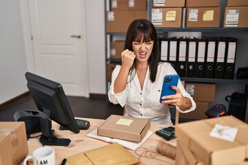 Young brunette woman working at small business ecommerce using smartphone annoyed and frustrated shouting with anger, yelling crazy with anger and hand raised