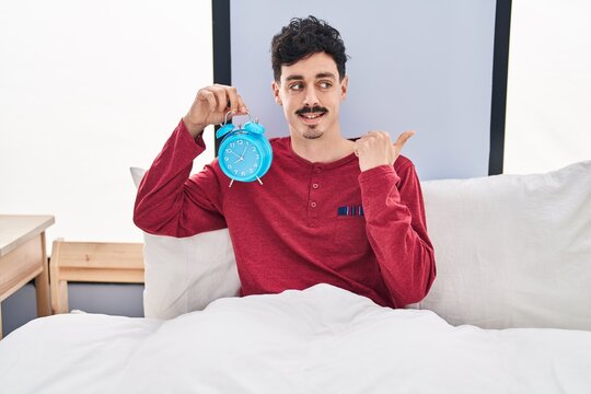 Hispanic man holding alarm clock in the bed pointing thumb up to the side smiling happy with open mouth