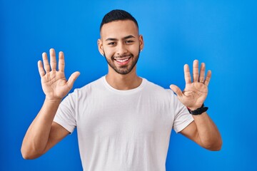 Young hispanic man standing over blue background showing and pointing up with fingers number ten while smiling confident and happy.