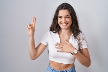 Young teenager girl standing over white background smiling swearing with hand on chest and fingers...