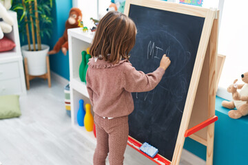 Adorable hispanic girl preschool student writing name on blackboard at kindergarten