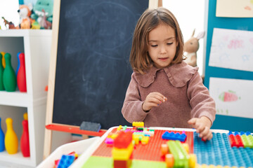 Adorable hispanic girl playing with construction blocks sitting on table at kindergarten
