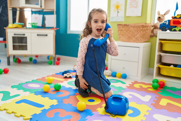 Adorable blonde girl playing with telephone toy sitting on floor at kindergarten - obrazy, fototapety, plakaty
