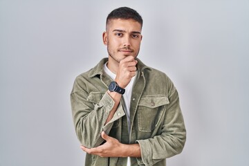 Young hispanic man standing over isolated background looking confident at the camera smiling with crossed arms and hand raised on chin. thinking positive.