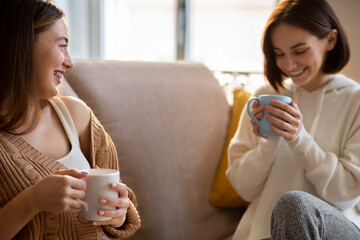 Laughing millennial european friends in sweaters with cups of coffee, sit on sofa enjoy...