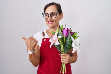 Middle age brunette woman wearing apron working at florist shop holding bouquet smiling with happy face looking and pointing to the side with thumb up.