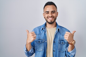 Young hispanic man standing over isolated background success sign doing positive gesture with hand, thumbs up smiling and happy. cheerful expression and winner gesture.