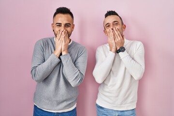 Homosexual couple standing over pink background laughing and embarrassed giggle covering mouth with hands, gossip and scandal concept