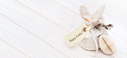 Easter bunny shaped Ivory gift bag with sweets and a gift tag with Happy Easter text and paper honeycomb Scandinavian craft Easter egg on white wooden table banner. Willow branch