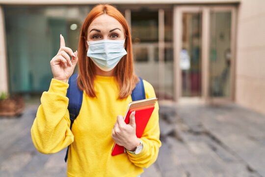 Young woman wearing safety mask and student backpack holding books surprised with an idea or question pointing finger with happy face, number one