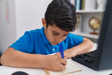 Adorable hispanic boy student using computer writing on notebook at classroom