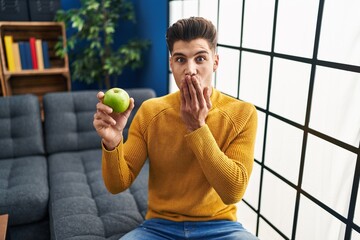 Young hispanic man holding green apple covering mouth with hand, shocked and afraid for mistake....
