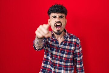 Young hispanic man with beard standing over red background pointing displeased and frustrated to...