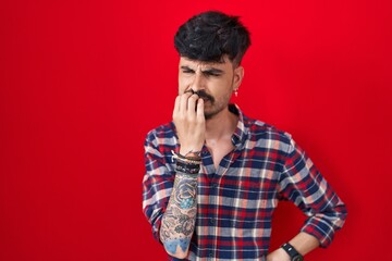 Fototapeta premium Young hispanic man with beard standing over red background looking stressed and nervous with hands on mouth biting nails. anxiety problem.