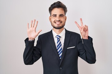 Young hispanic man with tattoos wearing business suit and tie showing and pointing up with fingers number seven while smiling confident and happy.