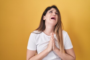 Young brunette woman standing over yellow background begging and praying with hands together with...