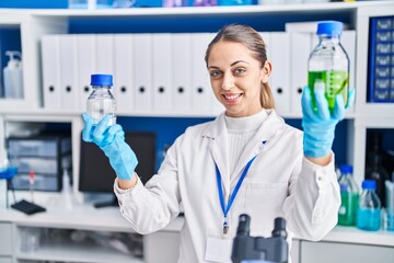 Young woman scientist smiling confident holding bottles with liquid at laboratory