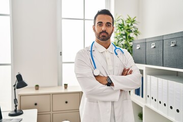 Young hispanic man wearing doctor uniform standing with arms crossed gesture at clinic