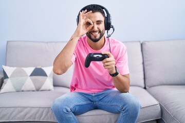 Fototapeta premium Hispanic young man playing video game holding controller sitting on the sofa doing ok gesture with hand smiling, eye looking through fingers with happy face.