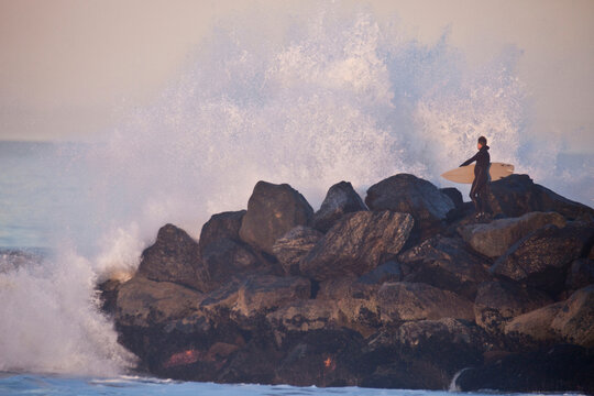 Surfer standing on a jetty checking out a large wave crashing over the end of the rocks.
