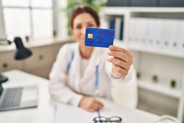 Young hispanic woman wearing doctor uniform holding credit card at clinic