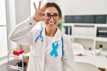 Young brunette doctor woman wearing stethoscope at the clinic doing ok gesture with hand smiling, eye looking through fingers with happy face.